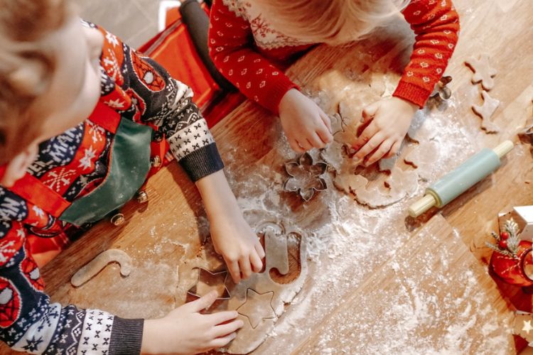 two young children in holiday sweaters making gingerbread men with cookie cutters