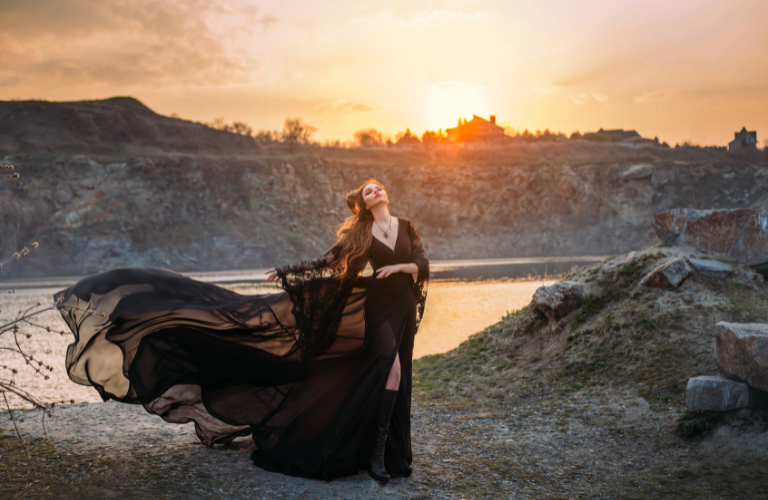 woman on a cliff with water and sunset behind her wearing a flowing black mesh dress