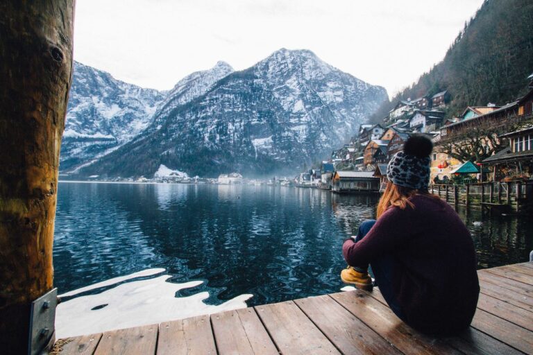 girl sitting on a dock looking across the lake at snowy mountains with a small village to the right